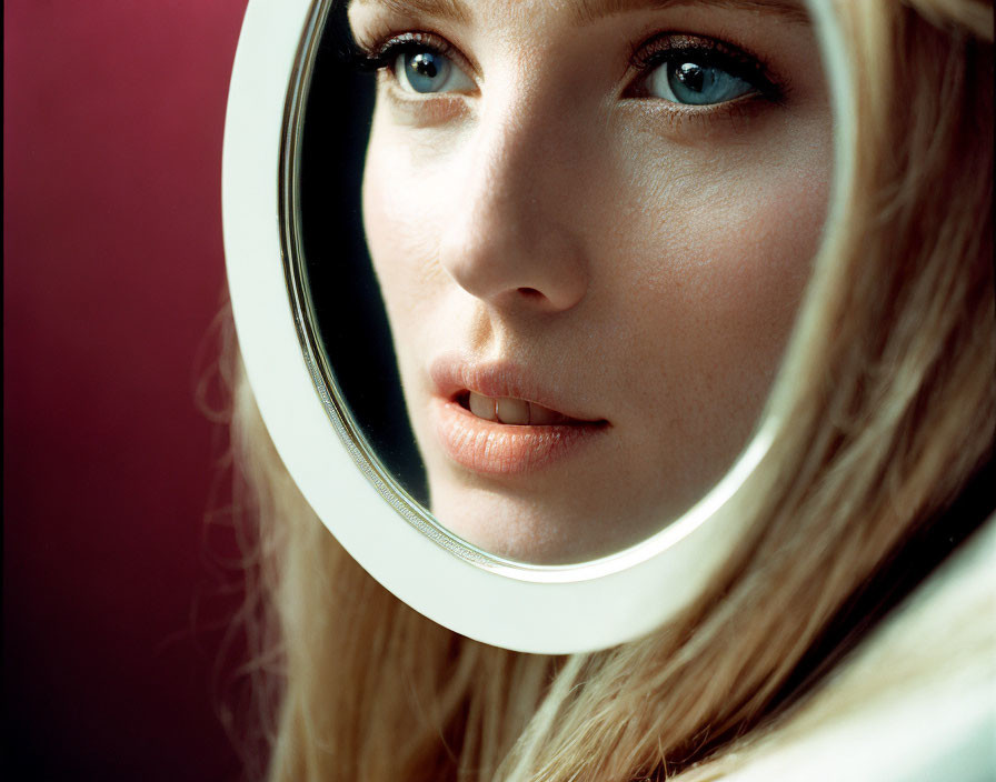 Woman with Blue Eyes Reflected in Round Mirror on Maroon Background