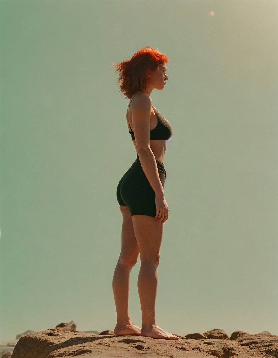 Red-haired woman in black swimsuit on sandy rock under clear sky