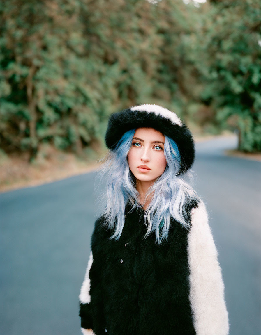 Blue-haired woman in black and white fur hat and vest on road with greenery