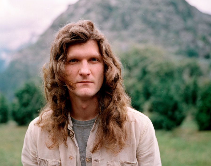 Long-haired man in casual attire against mountain backdrop