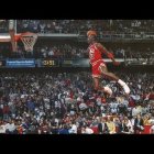 Intense basketball game with red-clad player mid-air for slam dunk, white players and crowd in