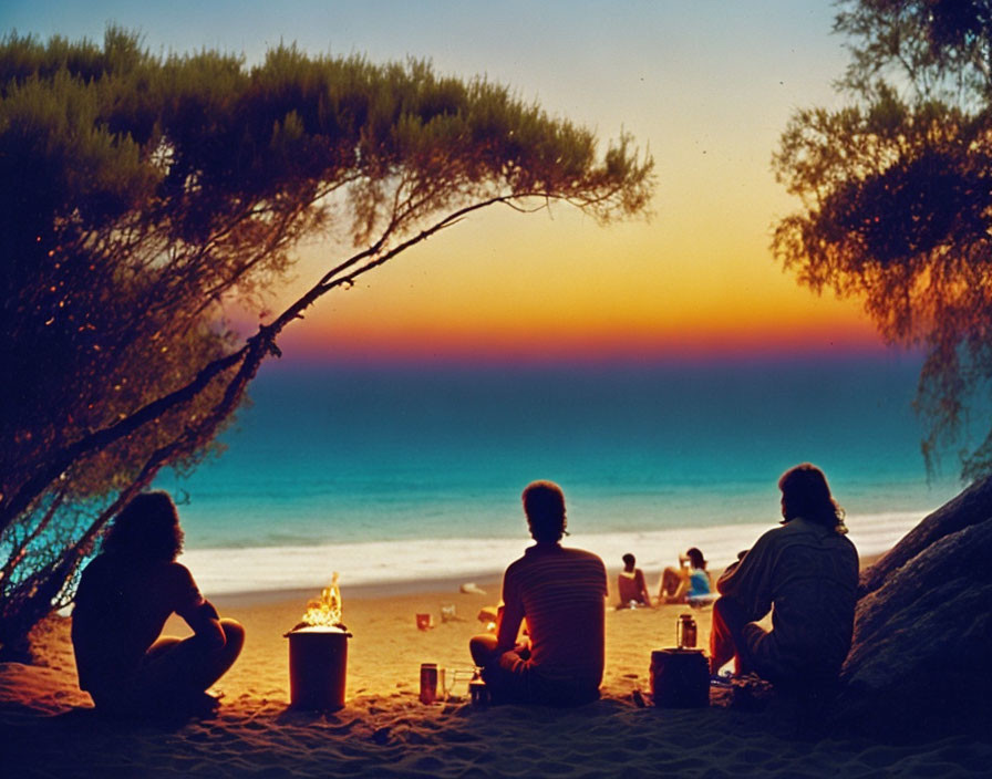 Three individuals by beach campfire at sunset with ocean view.