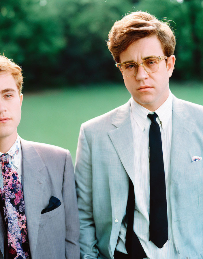 Two men in vintage suits outdoors with greenery backdrop.