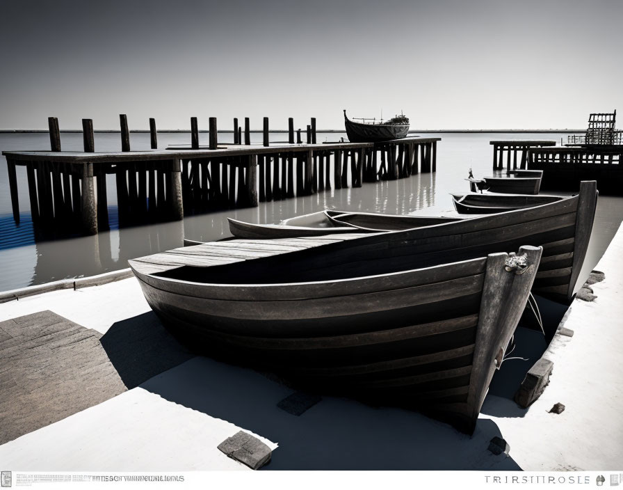 Monochrome photo of wooden rowboats on calm shore with dilapidated piers.