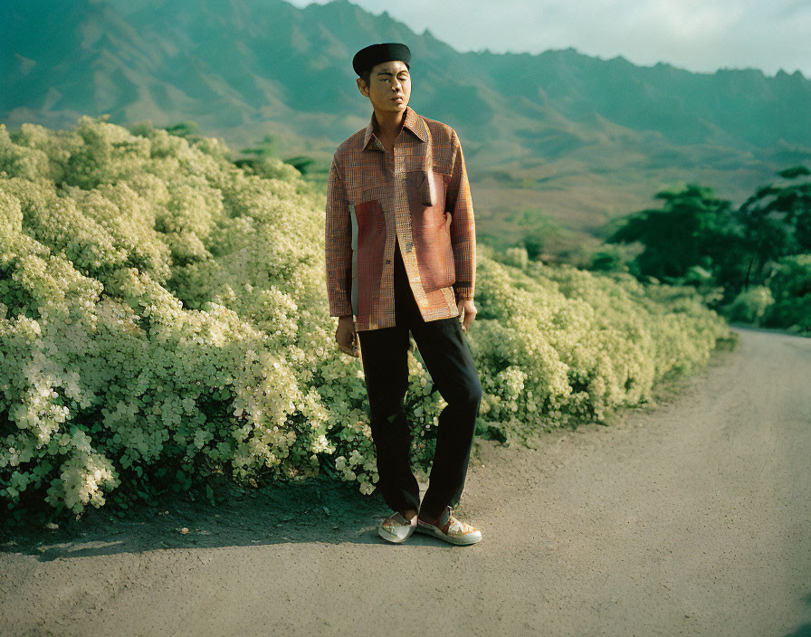 Person in plaid shirt and hat on dirt road with mountain backdrop