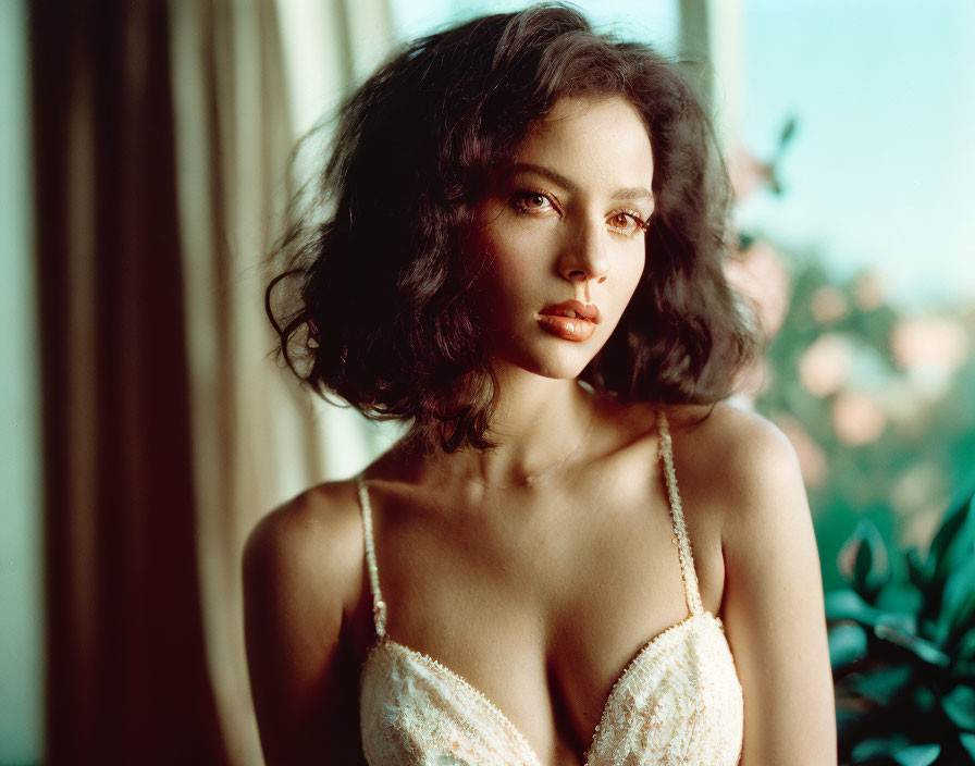 Curly-haired woman in white dress gazes by window with greenery.