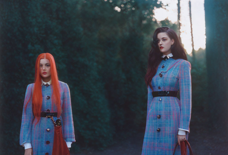 Two women in vintage plaid dresses in forest at dusk, one with red hair, one brunette.