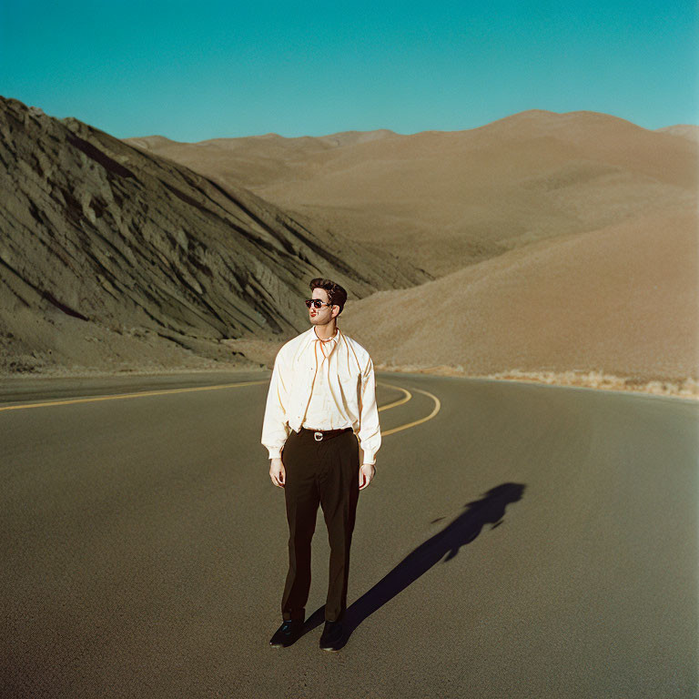 Man standing on curving desert road in sunglasses and white shirt.