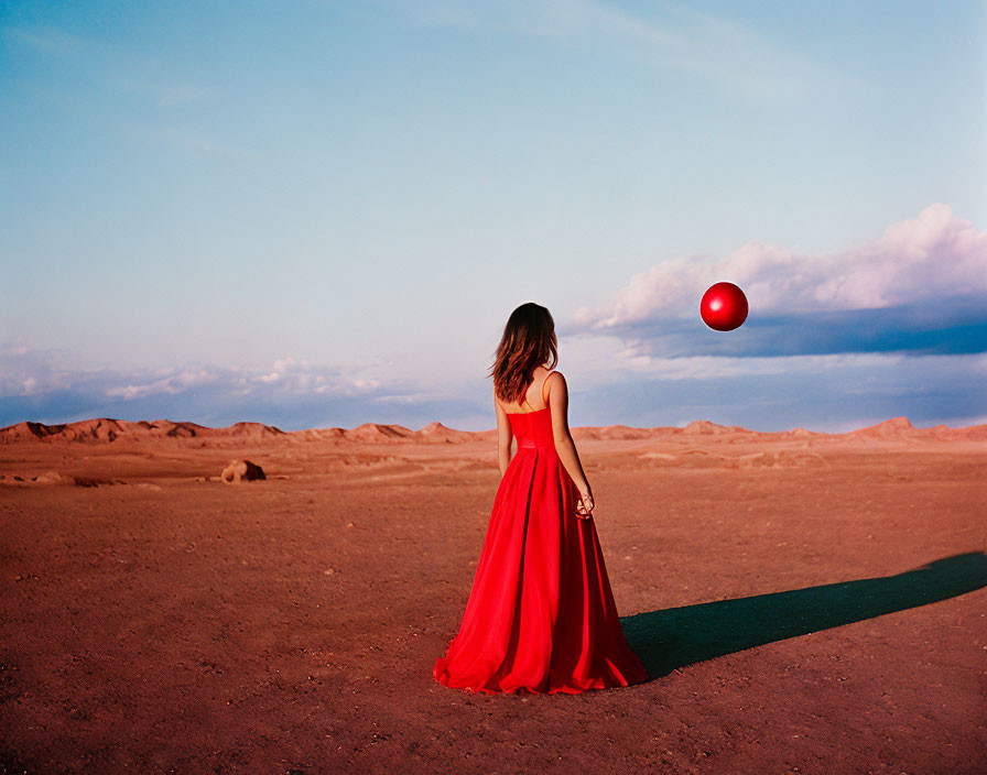 Woman in red dress gazes at floating red balloon in desert landscape