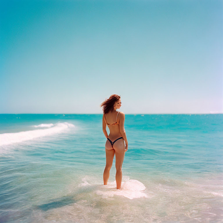 Person in one-piece swimsuit standing in clear sea water under blue sky