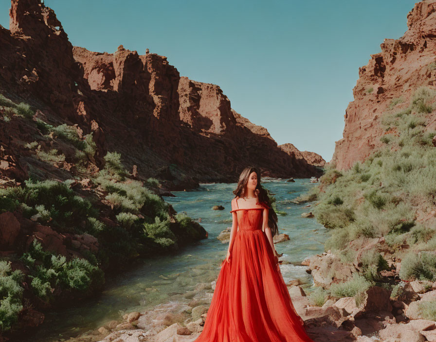 Woman in flowing red dress by river with cliffs and greenery