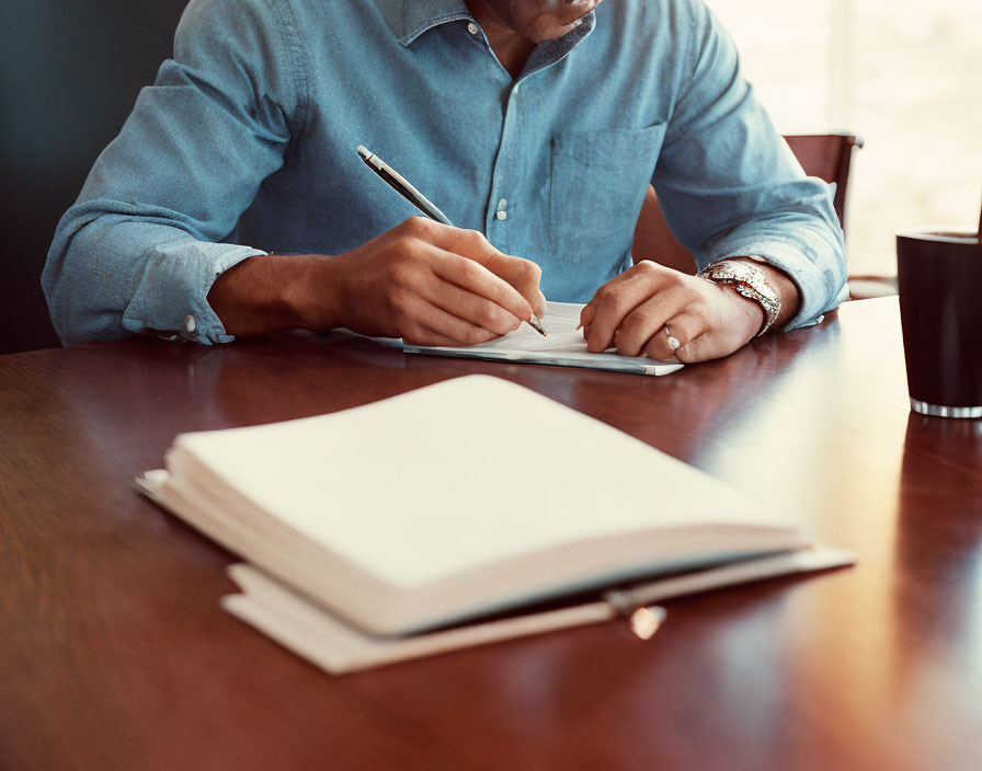 Person in Blue Shirt Writing in Notebook on Wooden Table with Open Book and Black Cup