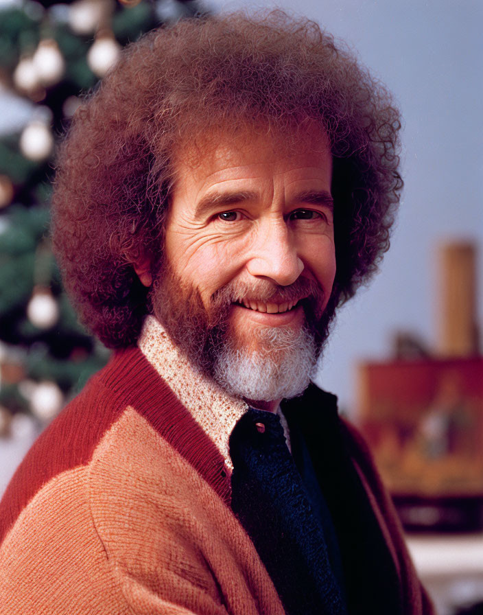 Smiling man with curly hair and beard in red-brown cardigan against greenery.