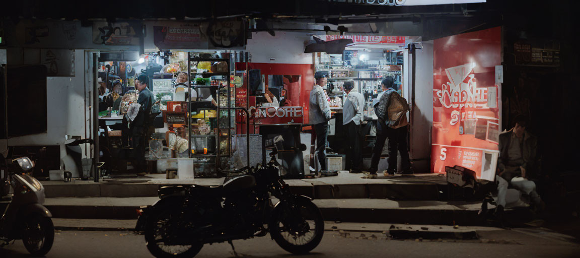 Night scene of a busy street shop with interior lights, customers, and parked motorcycle.