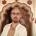 Man with Long Hair and Glasses in Front of Ornamental Wall wearing White Shirt and Necklace