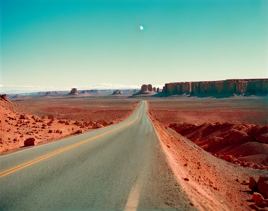 Desert road leading to rock formations under crescent moon on blue sky