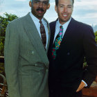 Two men in suits against cityscape: older with gray hair, younger with dark hair, both serious