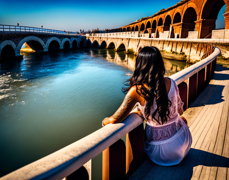 Woman with long dark hair in white top gazes at river from bridge under sunny sky