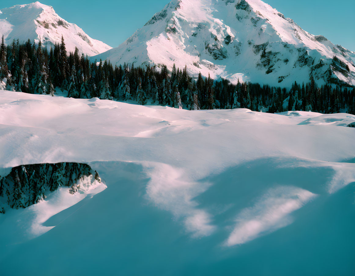 Snow-covered mountain landscape with sunlit peaks and forests under clear blue sky