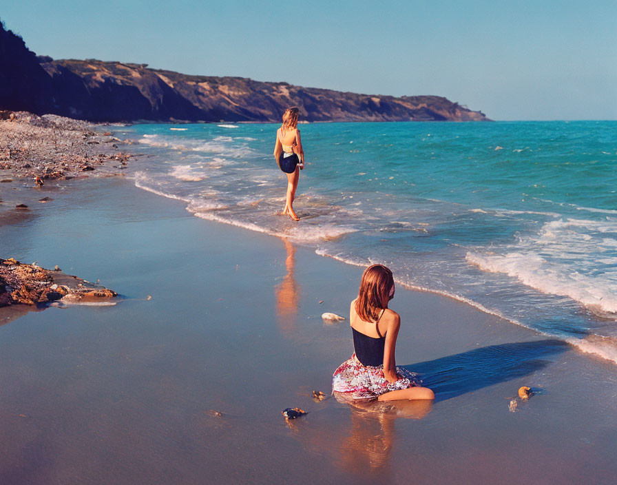 Two people at beach with one standing and one sitting, hills and clear sky in background.