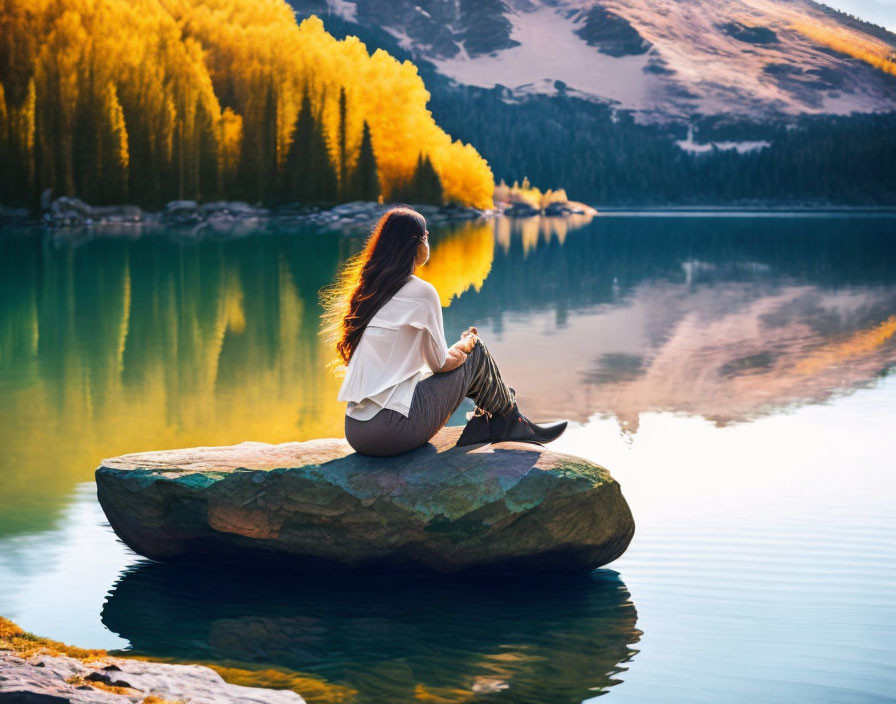 Person sitting by alpine lake with autumn trees and mountains reflected in water