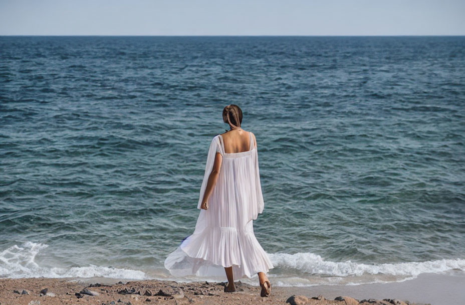 Woman in white dress walking on pebble beach towards sea