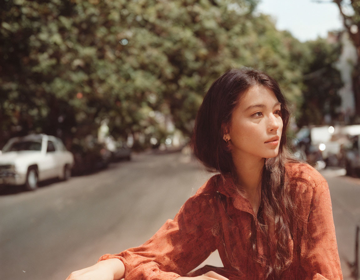 Pensive woman in orange shirt on sunny street with parked cars