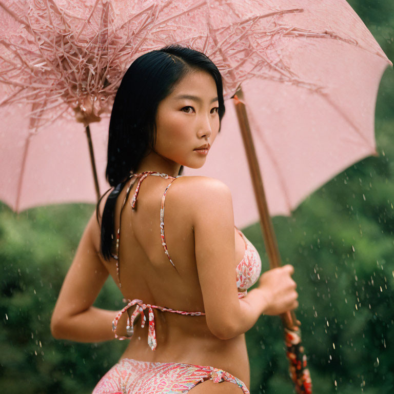 Woman with pink umbrella in rain, wearing patterned bikini amidst lush greenery