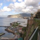 Coastal scene with boats, stone staircase, buildings, mountain backdrop, and cloud-shaped sky.