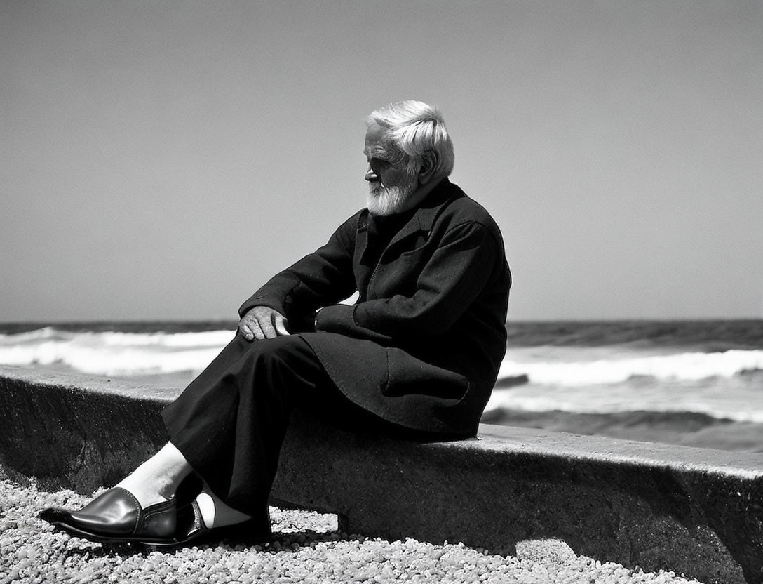 Elderly man with white beard in black attire sitting on coastal barrier.