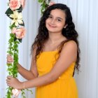 Young girl in yellow dress with long wavy hair holding green beads beside floral arrangement on white backdrop