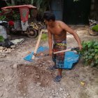 Person in hard hat and skirt digging near sand pile with universe pan, bulldozer in forest.