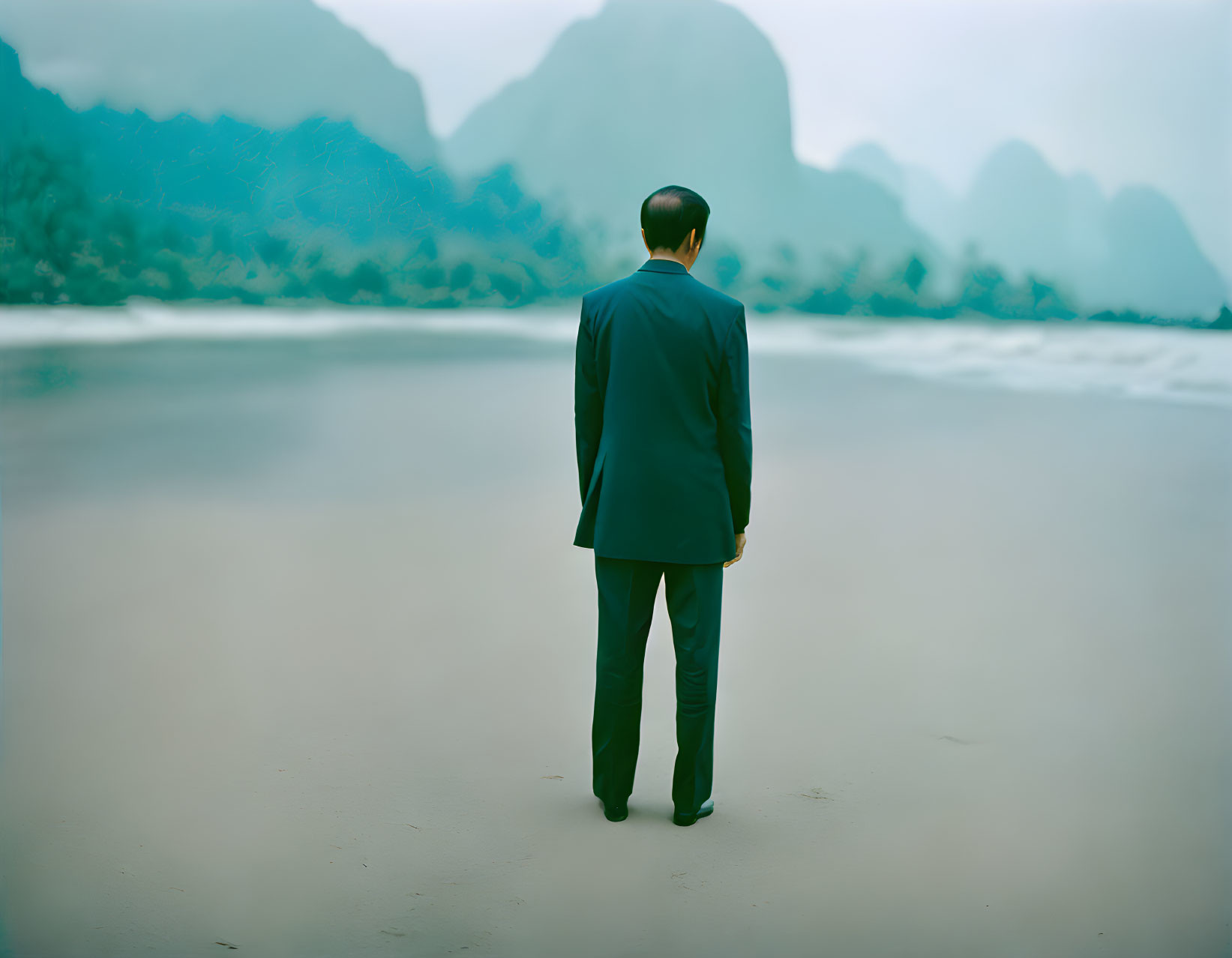 Man in suit by tranquil water with misty mountains and overcast sky