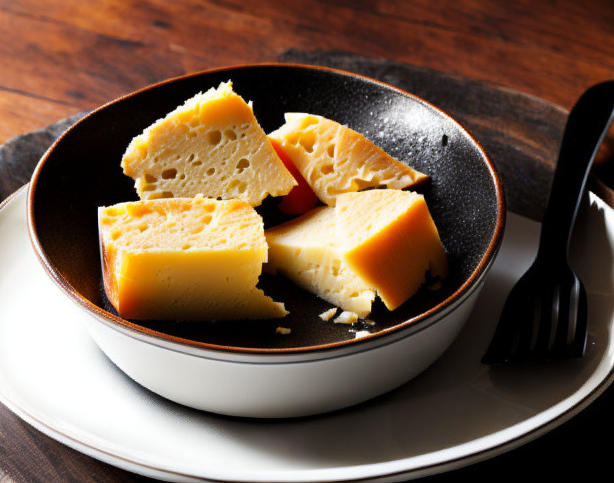 Porous yellow cheese slices on dark plate with fork on wooden table