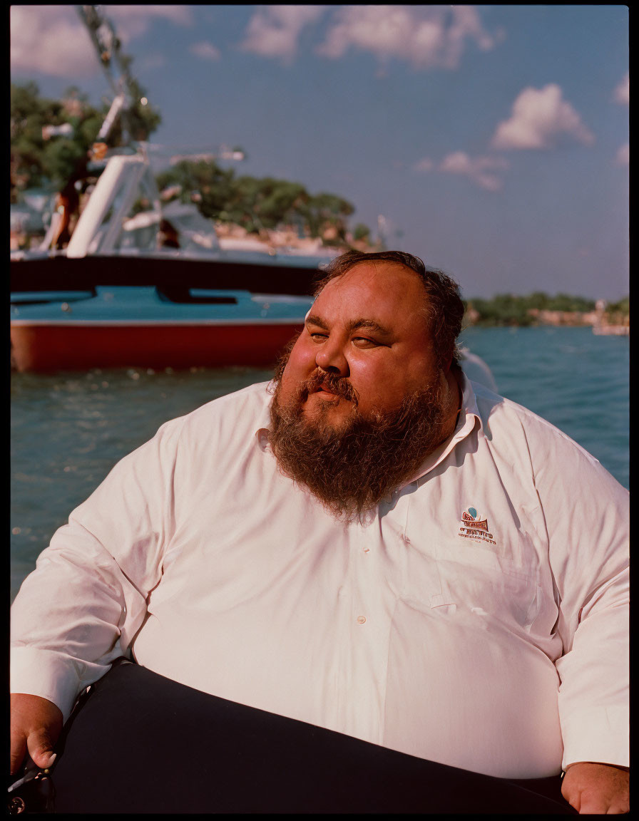 Bearded man in white shirt outdoors with sky, trees, boat