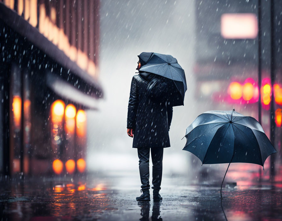 Person standing on rainy city street at night with umbrella and closed umbrella on wet pavement.