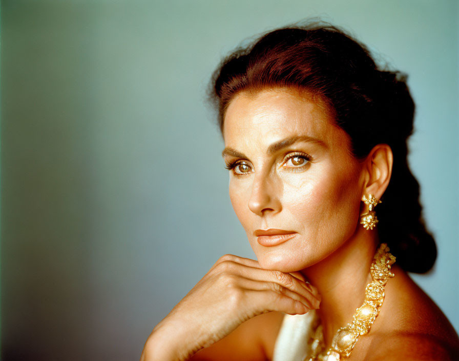 Dark-haired woman with striking features and jewelry posing against blue backdrop