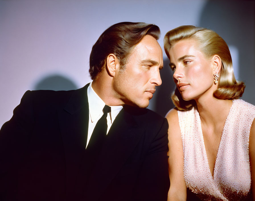 Man and woman in black suit and sparkling dress sitting closely with serious expressions under warm lighting