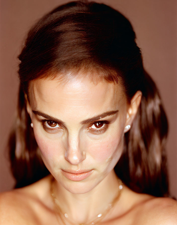 Dark-haired woman with intense gaze on warm-toned background wearing subtle jewelry