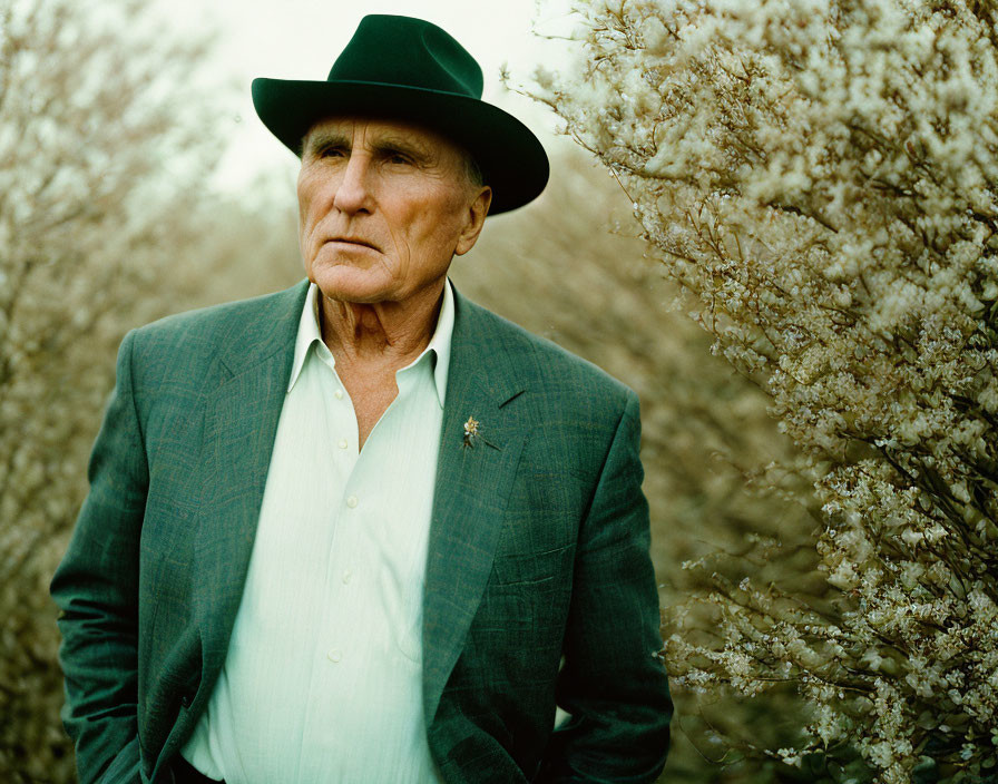 Elderly man in green suit and black hat gazes pensively at flowering shrubs