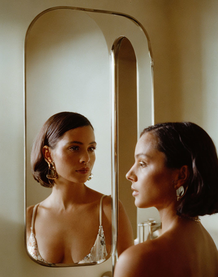 Woman admires reflection in vertical mirror wearing shimmering dress and elegant earrings against warm backdrop