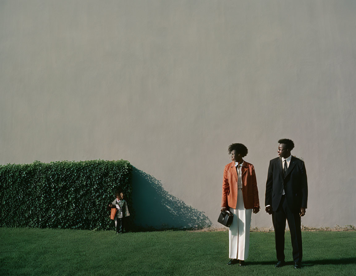 Elegantly dressed adults with child on grass against plain wall.