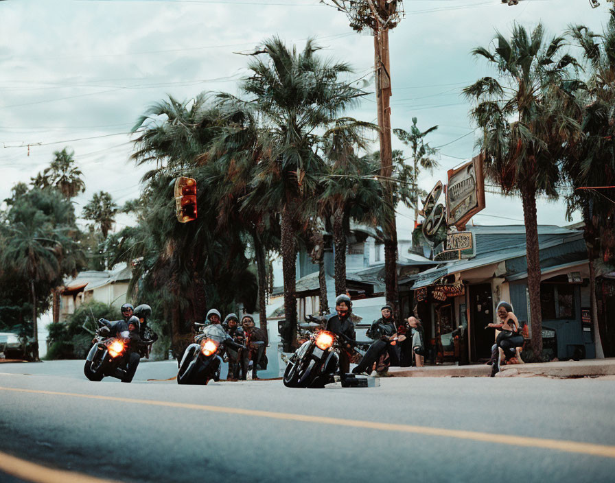 Motorcyclists riding in formation on street with palm trees and vintage storefronts.