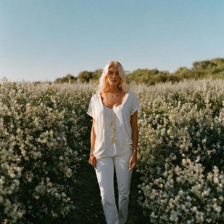 Woman in White Clothing Standing Among Blooming Bushes Under Clear Sky