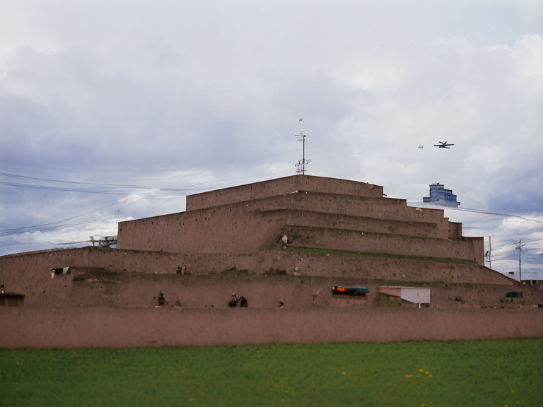 Terracotta terraced structure with grass and plane under cloudy sky