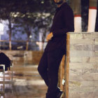 Man in black military-style outfit leaning against marble pillar in luxurious room with golden details.