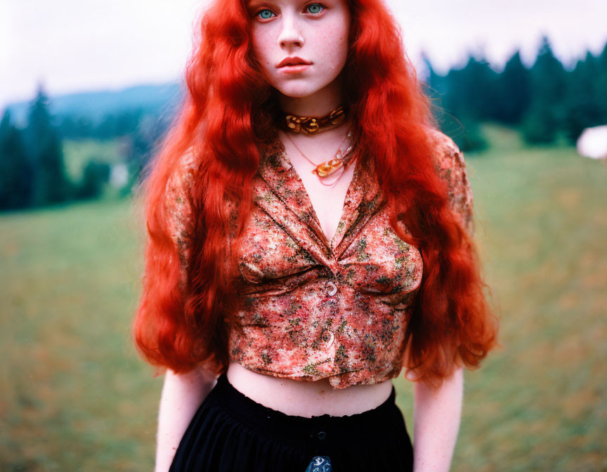 Red-haired woman in patterned blouse standing in grassy field with trees.