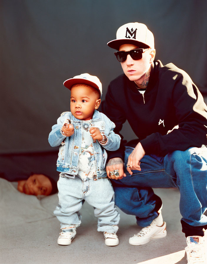 Toddler in denim jacket with adult in sunglasses and snapback next to person on ground.