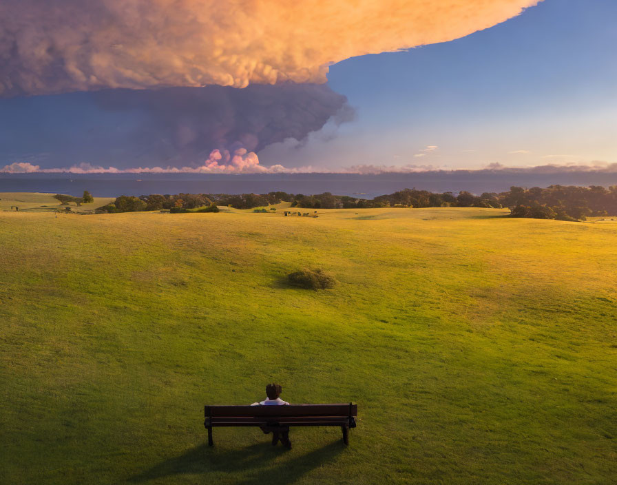 Person sitting on bench overlooking green field under dramatic sunset sky.