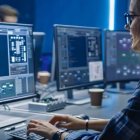 Person with glasses analyzes data on multiple computer monitors in dark room